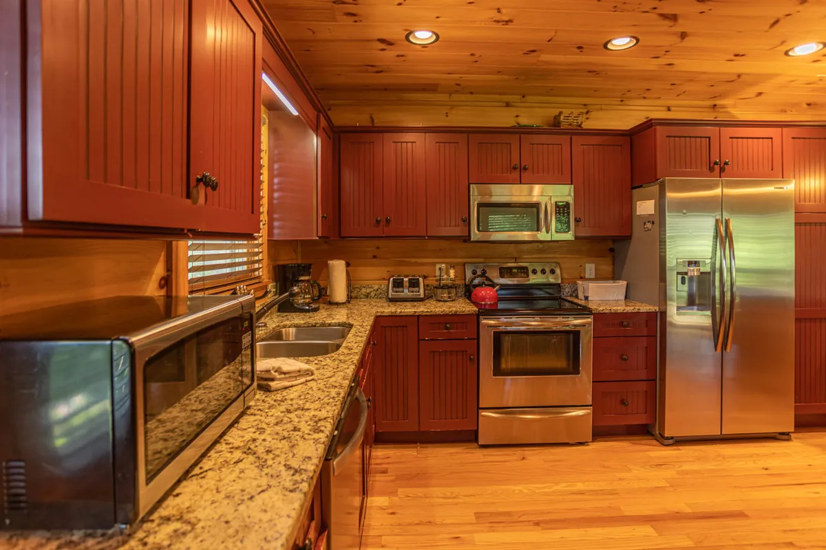 The image presents a kitchen with striking red cabinets, granite countertops, and modern stainless steel appliances. Warm wood paneling and spacious countertops provide the perfect setting for family gatherings and group meal prep.