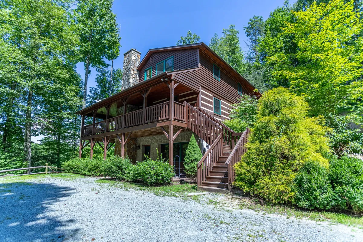 The image showcases the exterior of a beautiful log cabin nestled among lush greenery in the Blue Ridge Mountains. A spacious wooden deck wraps around the home, perfect for relaxing or entertaining, while a striking stone chimney hints at cozy evenings by the fire inside. The setting feels private and inviting, with staircases leading up to the deck and a backdrop of mature trees.