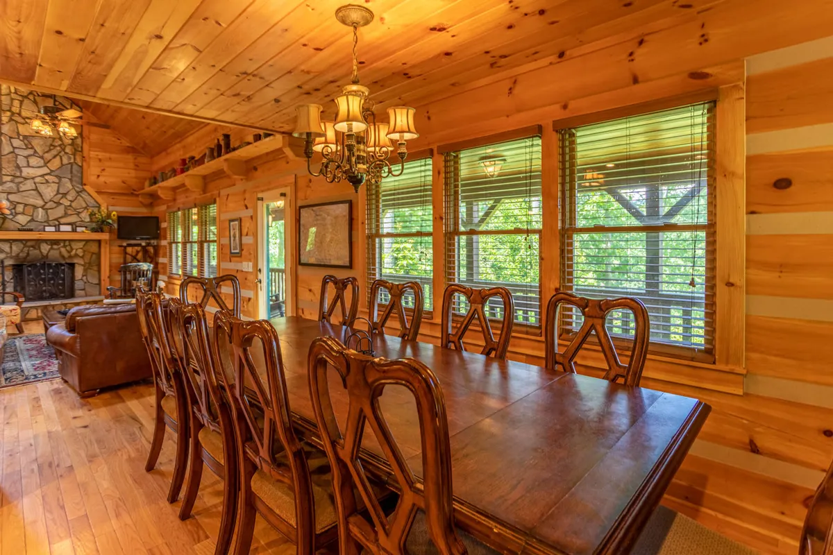 An elegant dining area with a substantial wooden table, glowing chandelier, stone fireplace, exposed wooden beams, and unobstructed mountain views visible through the windows. The atmosphere is both spacious and intimate, perfect for shared meals and celebrations.
