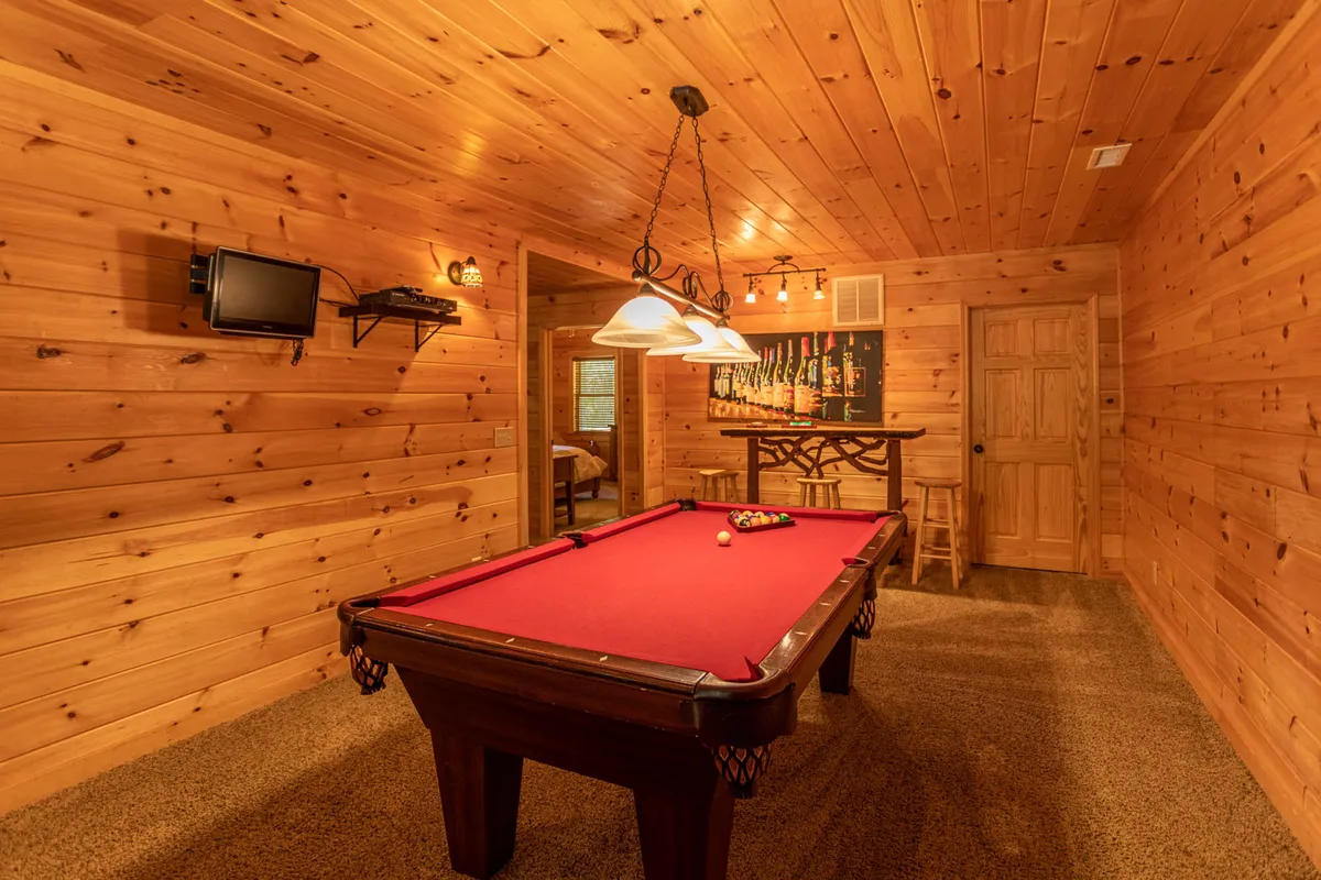 A game room featuring a pool table, light fixtures, warm wood paneling, and a bar area. The space looks inviting for group activities, encouraging friendly competition and casual socializing.