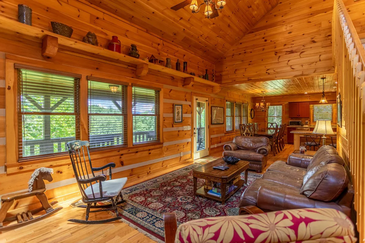 A sunlit living area featuring a rocking chair, wooden coffee table, and sofa set. Large windows frame scenic views, and the overall ambiance is relaxed and inviting—ideal for leisurely gatherings or quiet moments.