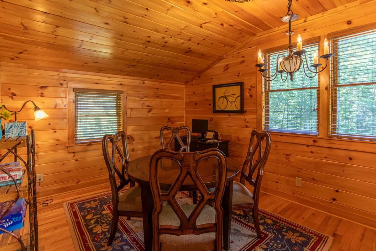 An upstairs loft area with a wooden dining table, chandelier, bookshelf, and windows. The setting is versatile for playing board games, working, or casual gatherings, echoing the section's depiction of a hub for games, crafts, and remote check-ins.