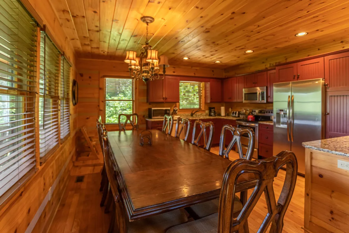 A large wooden dining table sits beneath a chandelier, surrounded by red cabinetry and expansive granite countertops. The space is designed for communal dining, perfect for game nights or family celebrations.