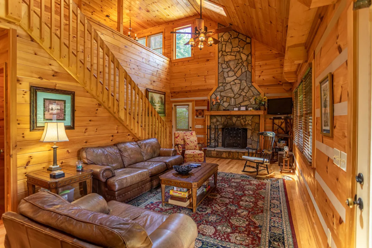 A warm, welcoming living room with a prominent stone fireplace as the centerpiece, flanked by leather sofas atop a handsome rug. Sunlight from large windows brightens the space, accentuating the rustic wooden staircase and beams above.