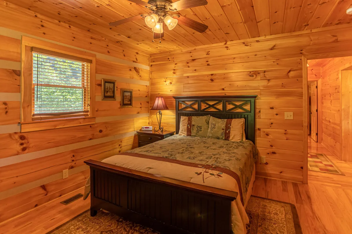 A warmly lit queen bedroom featuring a classic wooden bed frame with floral bedding, soft lampshade lighting, wood-paneled walls, and a window with blinds that lets in natural light. The cozy atmosphere is perfect for restful nights and peaceful mornings.