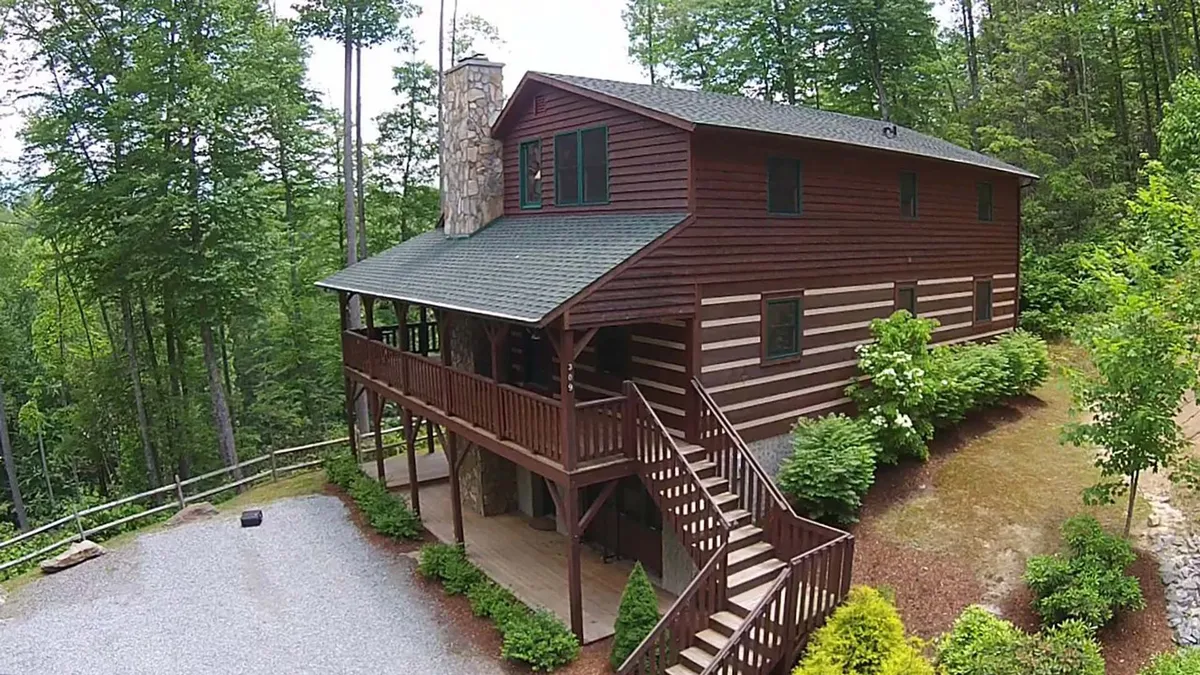 This image captures the exterior of the log cabin, showcasing its classic log construction, stone chimney, wooden stairs, and porch area surrounded by lush greenery. The scene exudes a welcoming and serene mountain retreat vibe, emphasizing the property's inviting outdoor spaces ideal for group gatherings and relaxation.
