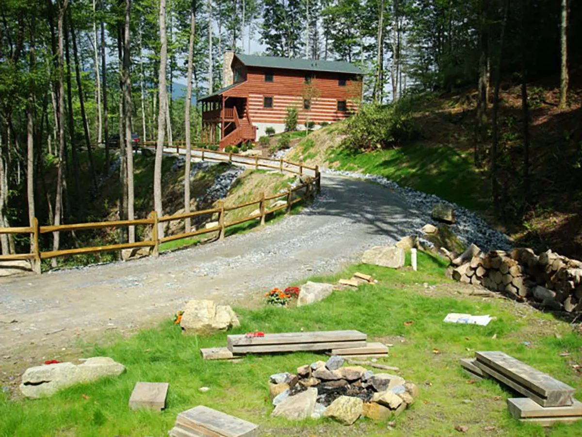 A wide exterior view of the log cabin nestled among trees, featuring the winding gravel driveway, spacious lawn, and a welcoming fire pit area with chairs. The scene showcases the property's outdoor amenities, inviting gathering spaces, and the tranquil mountain atmosphere.
