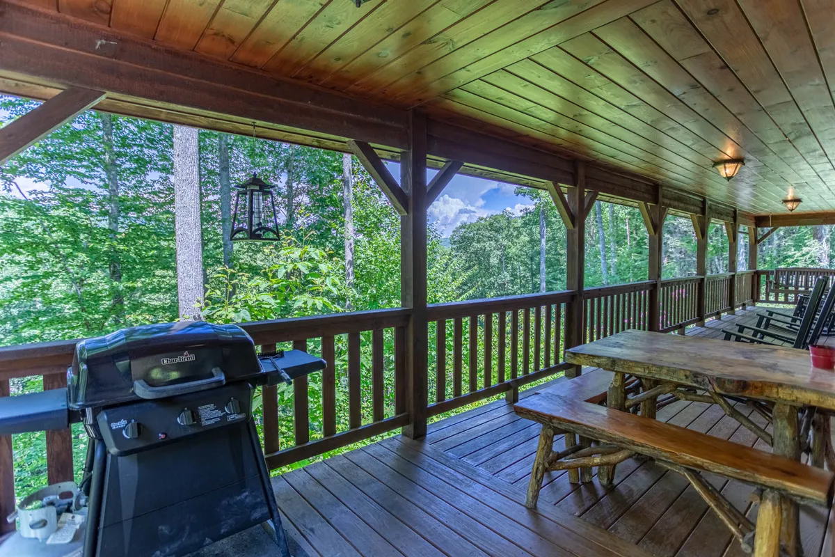 A covered deck outfitted with a grill, wooden table for outdoor dining, classic rocking chairs, and surrounded by lush greenery. Lanterns provide cozy ambient lighting, inviting guests to linger outside.